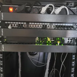Widok z bliska szafy rack z zamontowanymi patch panelami Lanberg, przełącznikiem Cisco SG200-26 z aktywnymi portami Gigabit Ethernet oznaczonymi zielonymi diodami oraz przewodami sieciowymi.