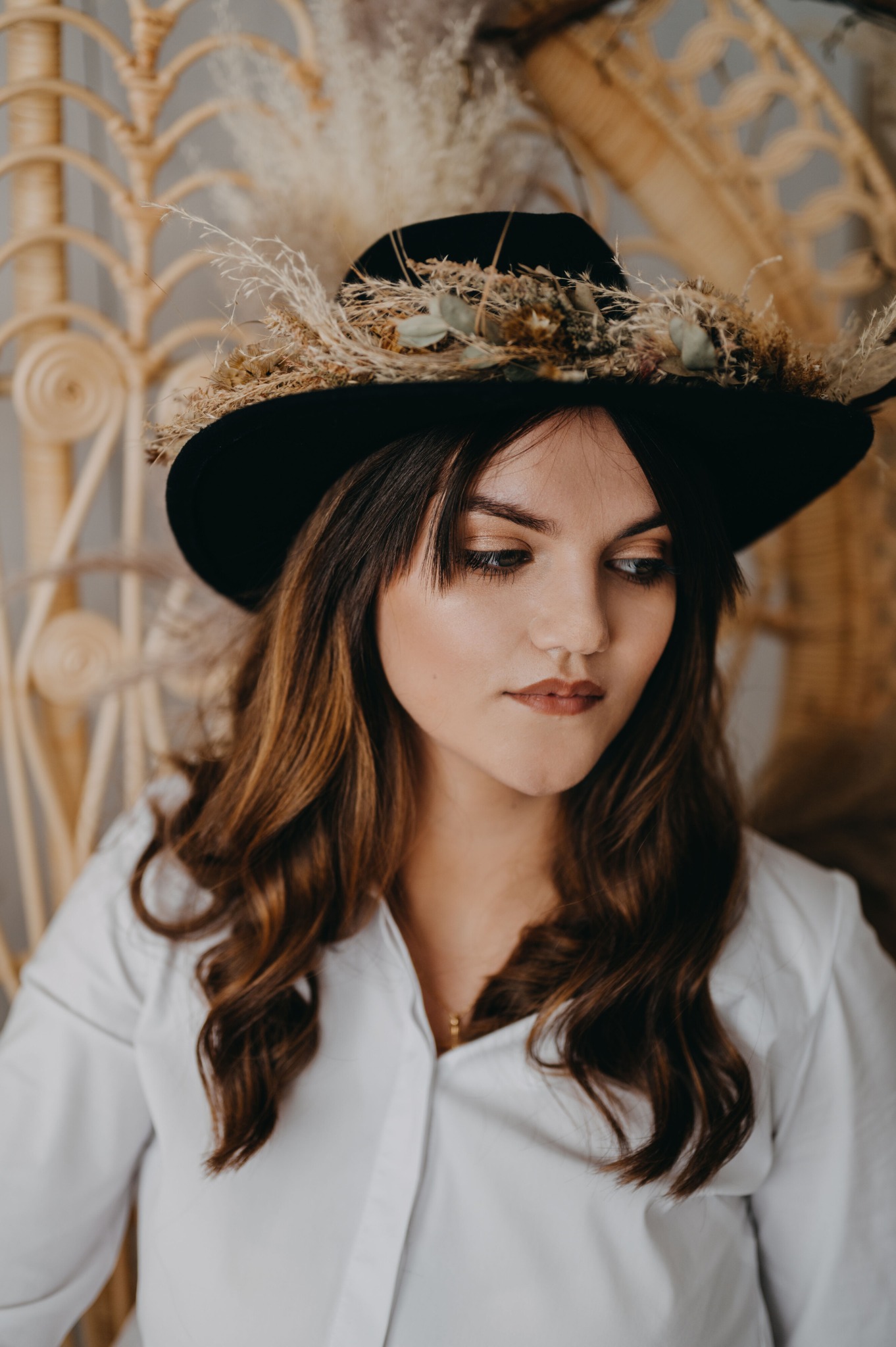 Portret kobiety z delikatnym makijażem, brązowymi włosami i czarnym kapeluszem ozdobionym suszonymi kwiatami, pozującej na tle wiklinowego krzesła.