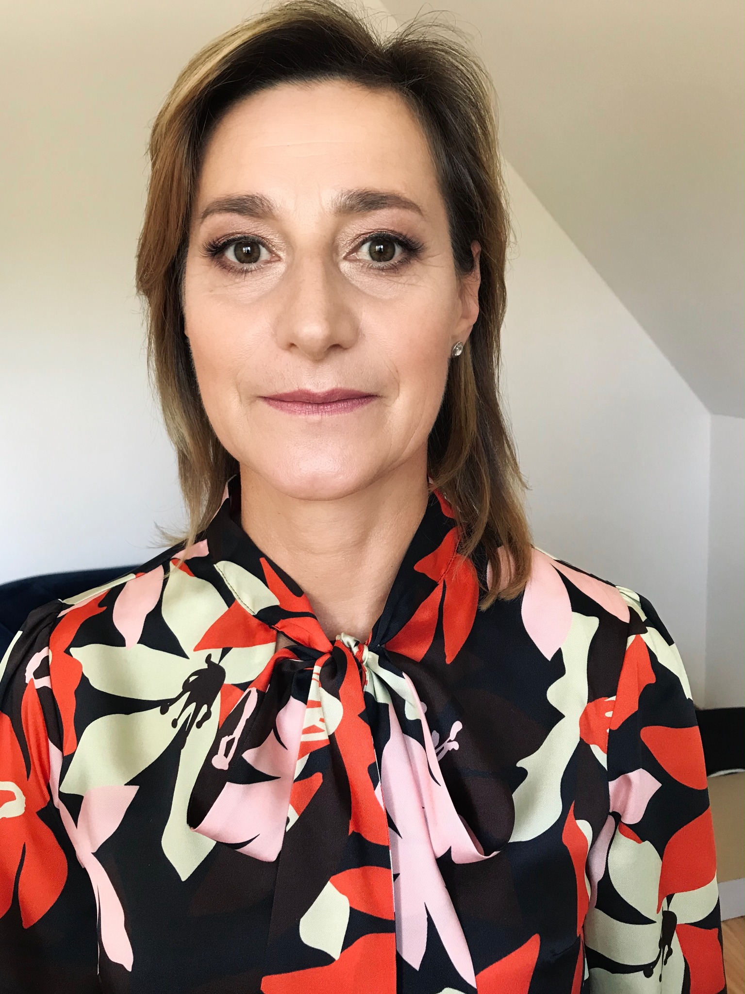 Portret kobiety z delikatnym makijażem oczu i ust, ubrana w kolorową bluzkę w kwiaty, z kolczykiem w uchu.