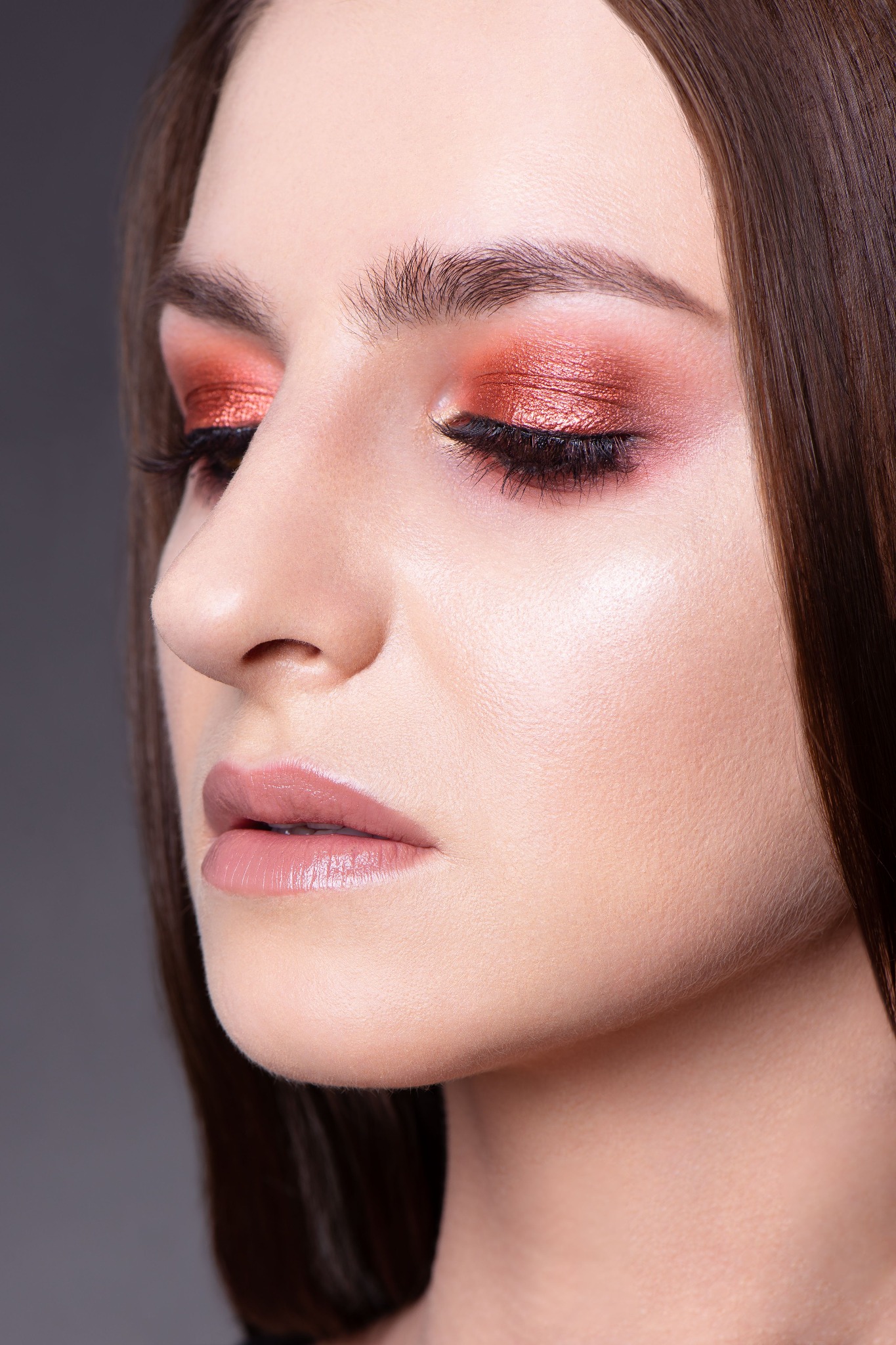 Profil kobiety z intensywnym, błyszczącym, pomarańczowo-czerwonym cieniem na powiekach i delikatnym, różowym błyszczykiem na ustach; gładka, rozświetlona cera.