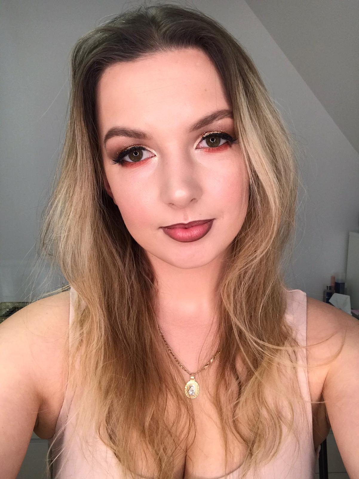 Portret kobiety z intensywnym makijażem oczu w odcieniach czerwieni i złota, podkreślonymi czarną kreską i długimi rzęsami, oraz z ciemną pomadką na ustach. Na szyi złoty łańcuszek z medalikiem.