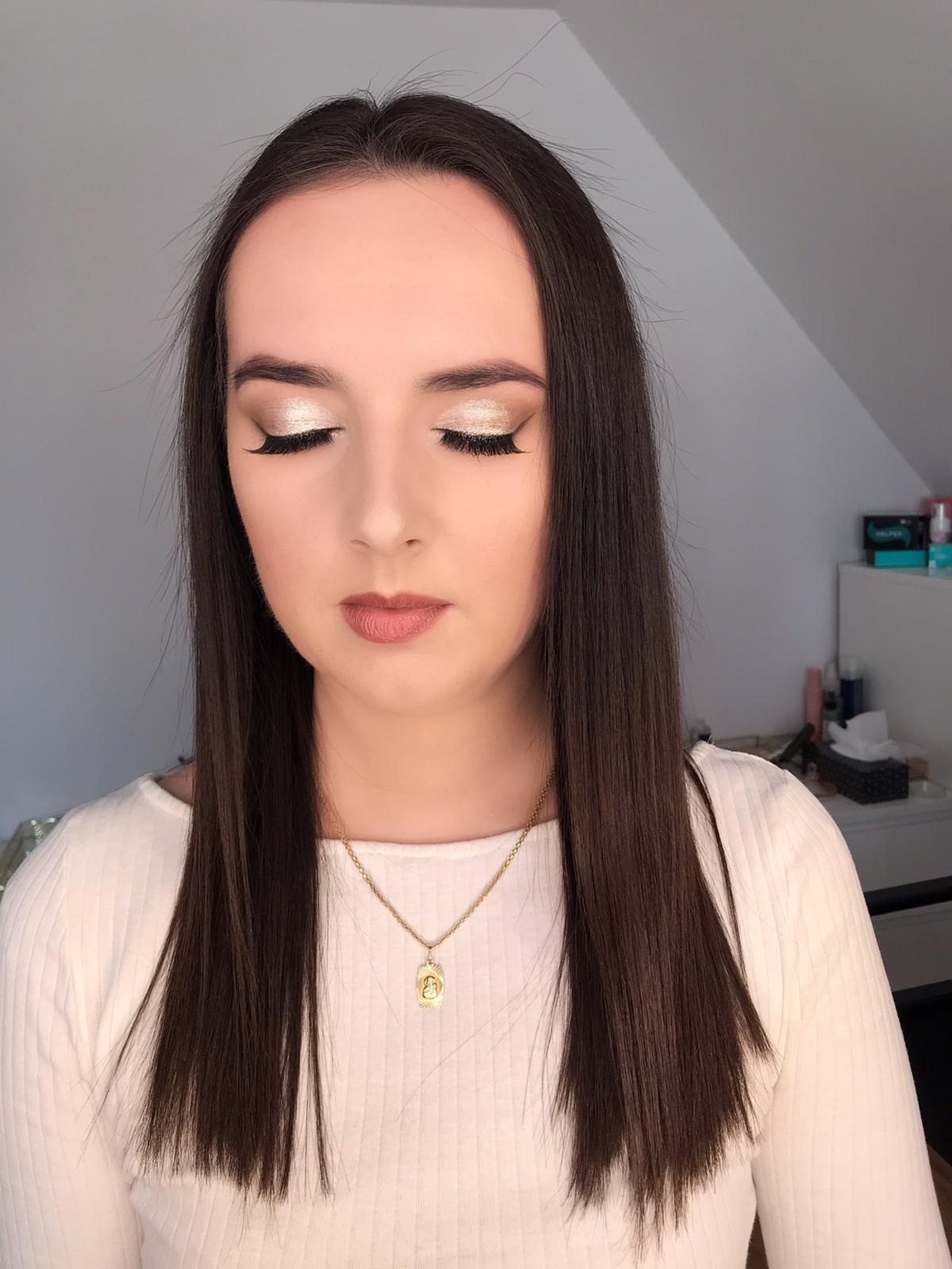 Portret kobiety z zamkniętymi oczami prezentującej złoty, błyszczący efekt na powiekach i delikatny makijaż ust, wykonany w Nowym Targu. Widoczny złoty naszyjnik i proste, ciemne włosy.