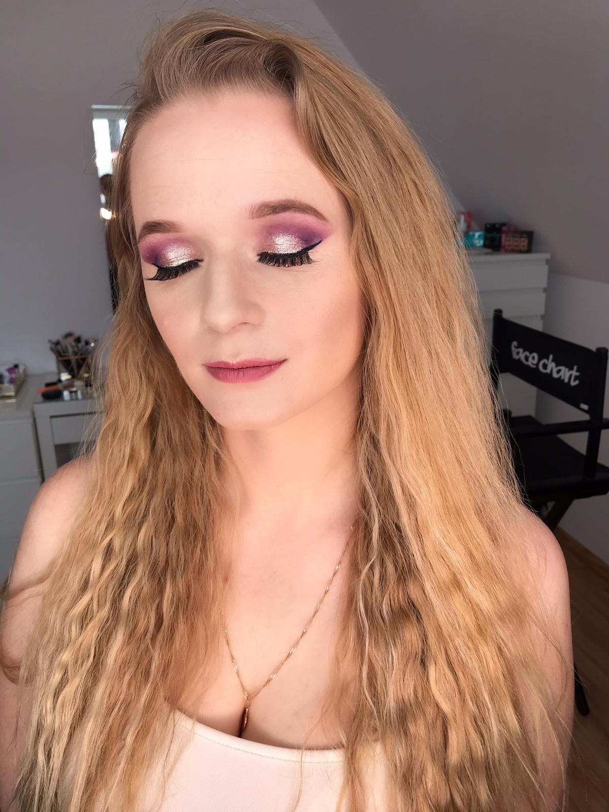 Portret kobiety z zamkniętymi oczami prezentującej fioletowo-różowy makijaż oczu z czarną kreską i sztucznymi rzęsami, długie blond włosy opadają na ramiona, widoczny złoty łańcuszek na szyi.