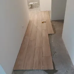 Układanie podłogi z paneli imitujących drewno w korytarzu, widoczne plastikowe krzyżyki dystansowe i miarka na ułożonej części podłogi, surowy beton na nieukończonej powierzchni.