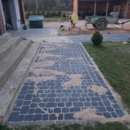 Ułożona, ciemna kostka brukowa przed schodami wejściowymi domu, częściowo zasypana piaskiem, w tle widoczny pracownik z zagęszczarką i przyczepą.