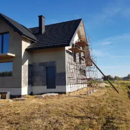 Dom w trakcie ocieplania, widoczne szare bloczki izolacyjne na ścianie, rusztowanie i wykop w ziemi, błękitne niebo w tle.
