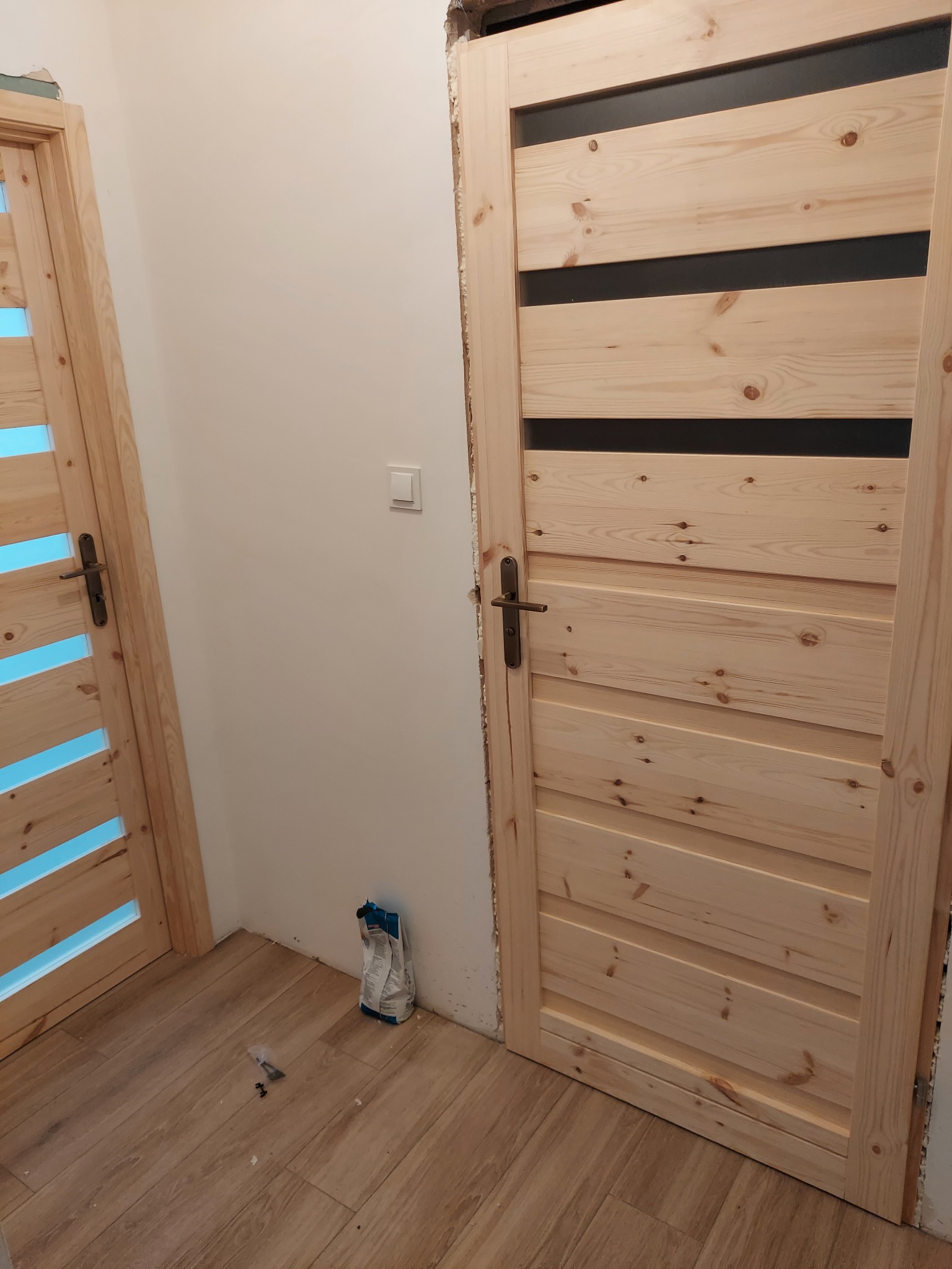 Jasne, drewniane drzwi wewnętrzne z poziomymi, czarnymi wstawkami, zamontowane w surowym otworze, obok drugich drzwi z przeszkleniami, na podłodze panele i worek gipsu.