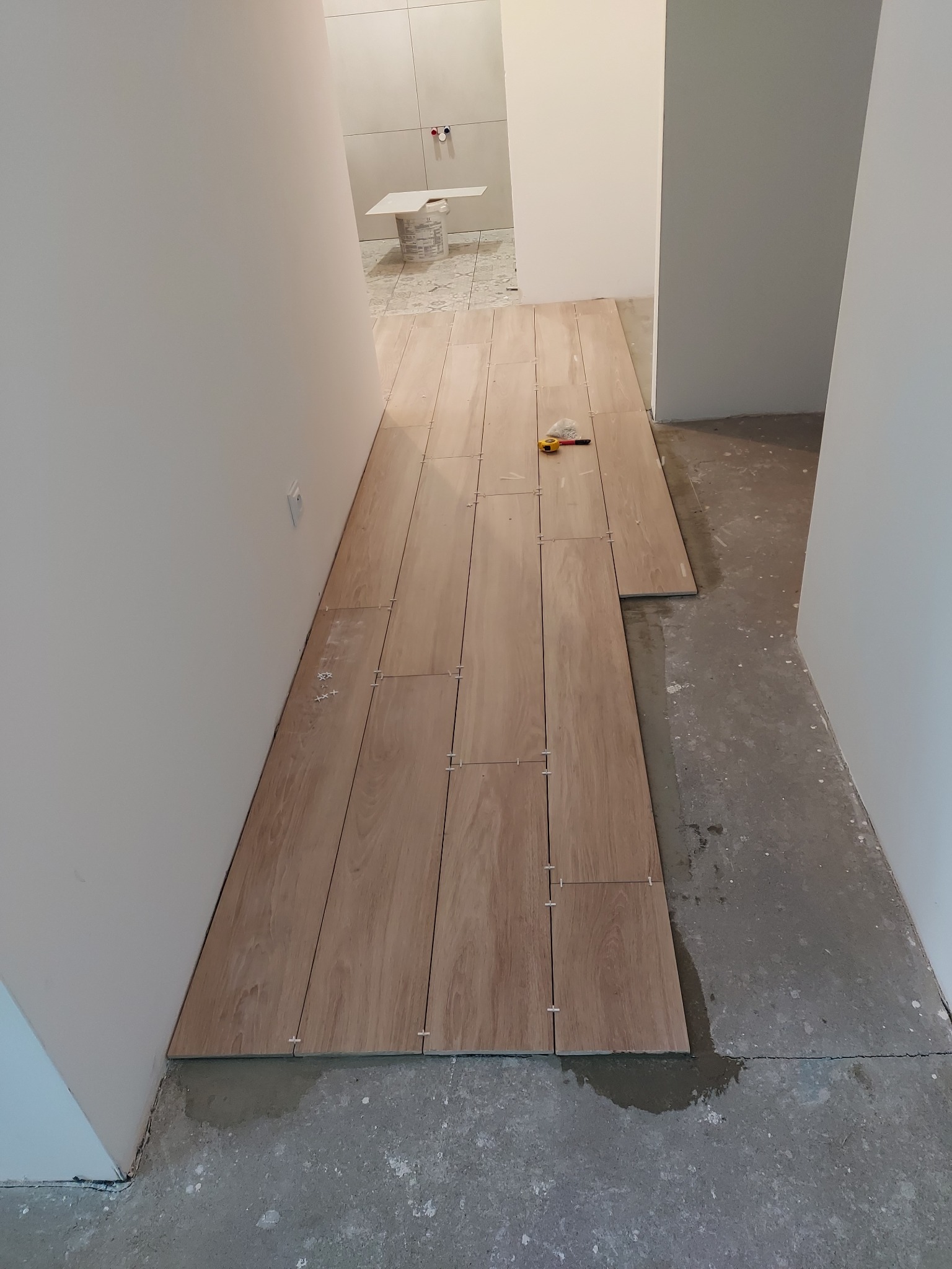 Układanie podłogi z paneli imitujących drewno w korytarzu, widoczne plastikowe krzyżyki dystansowe i miarka na ułożonej części podłogi, surowy beton na nieukończonej powierzchni.