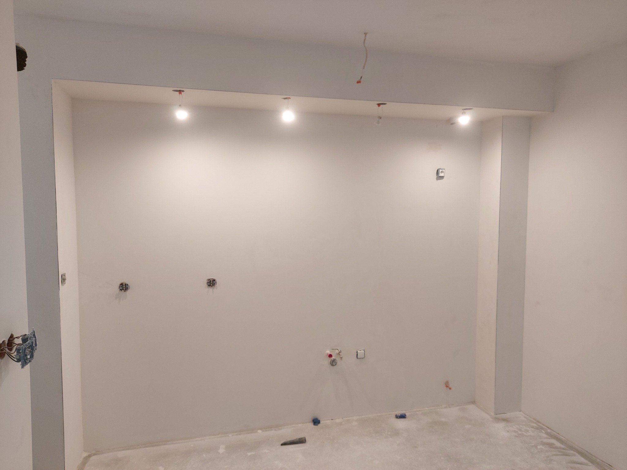 Przygotowana do wykończenia kuchnia z widocznymi instalacjami elektrycznymi i hydraulicznymi oraz świeżo pomalowanymi ścianami na biało i wbudowanymi punktowymi lampami sufitowymi.