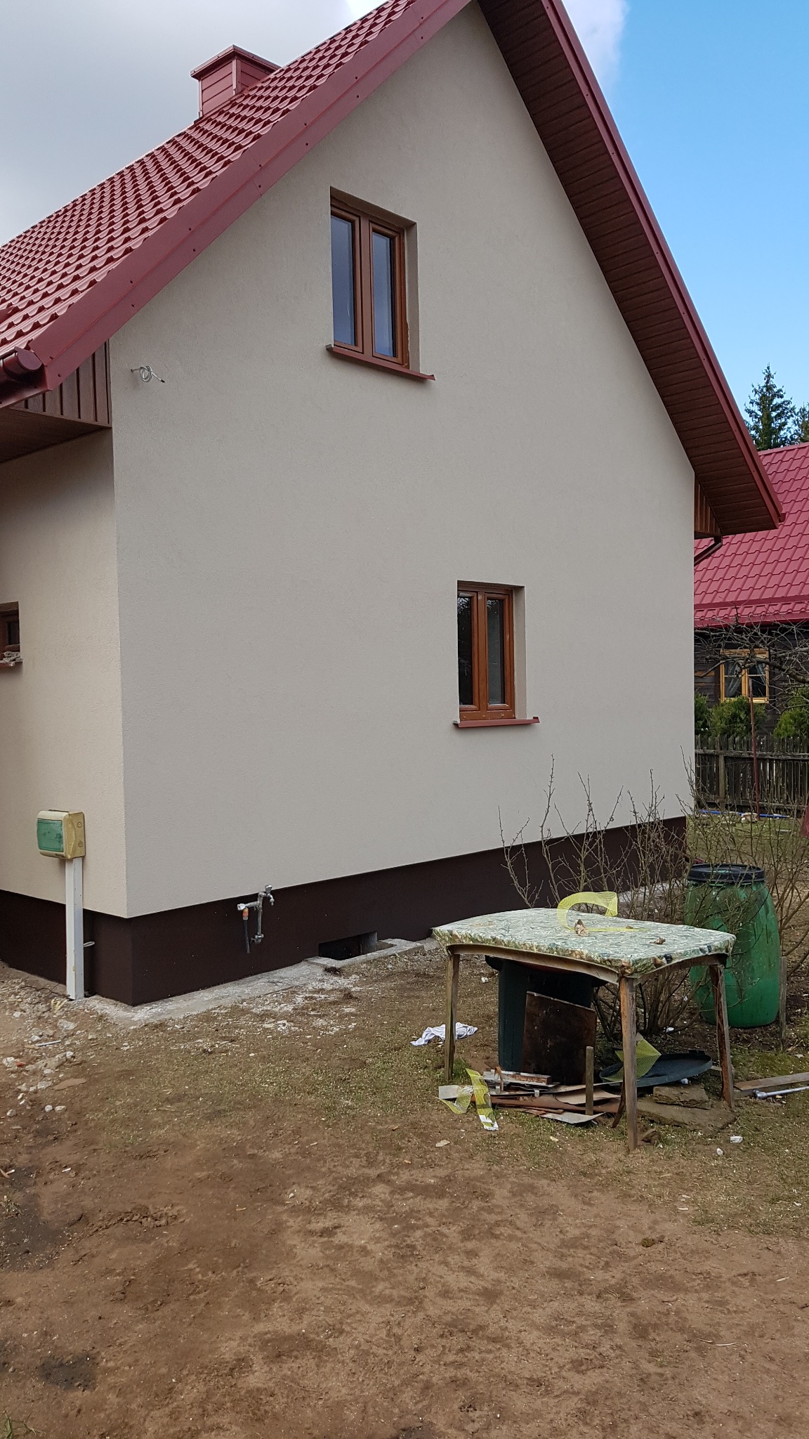 Świeżo otynkowana ściana domu jednorodzinnego z brązowymi oknami i czerwoną dachówką, widoczny cokół w kolorze brązowym, na trawniku prowizoryczny stół roboczy i nieporządek po pracach.