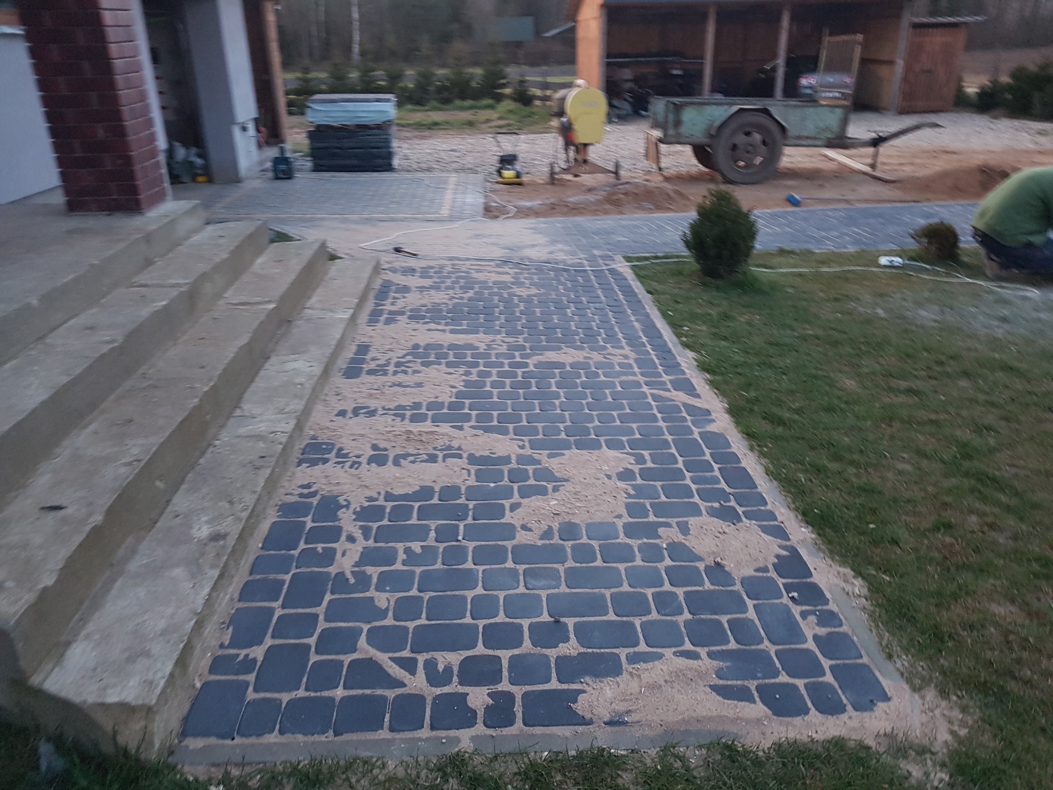 Ułożona, ciemna kostka brukowa przed schodami wejściowymi domu, częściowo zasypana piaskiem, w tle widoczny pracownik z zagęszczarką i przyczepą.