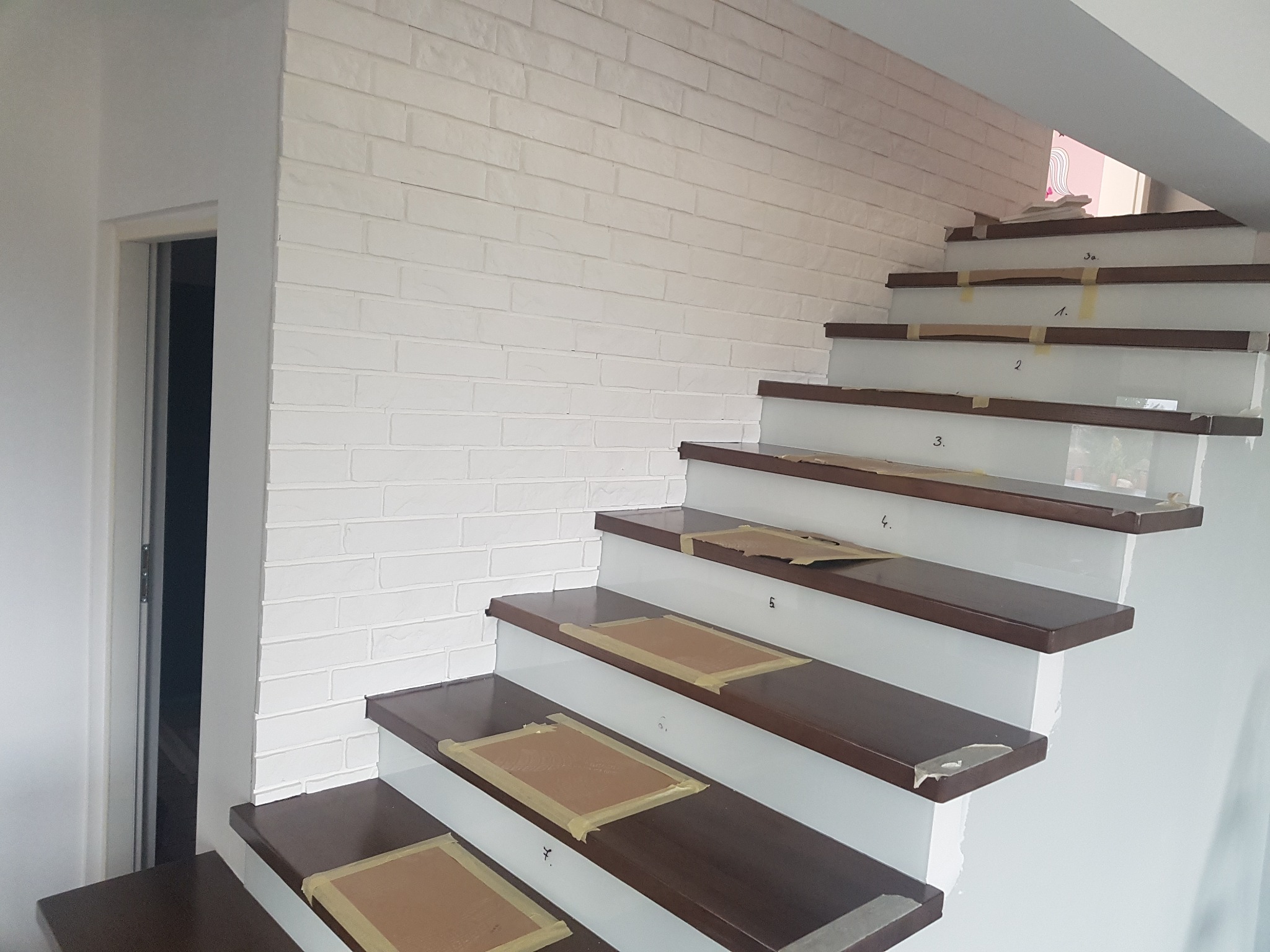 Montaż drewnianych stopni schodów z białymi podstopnicami, zabezpieczonych taśmą malarską, na tle ściany wykończonej białą cegłą.