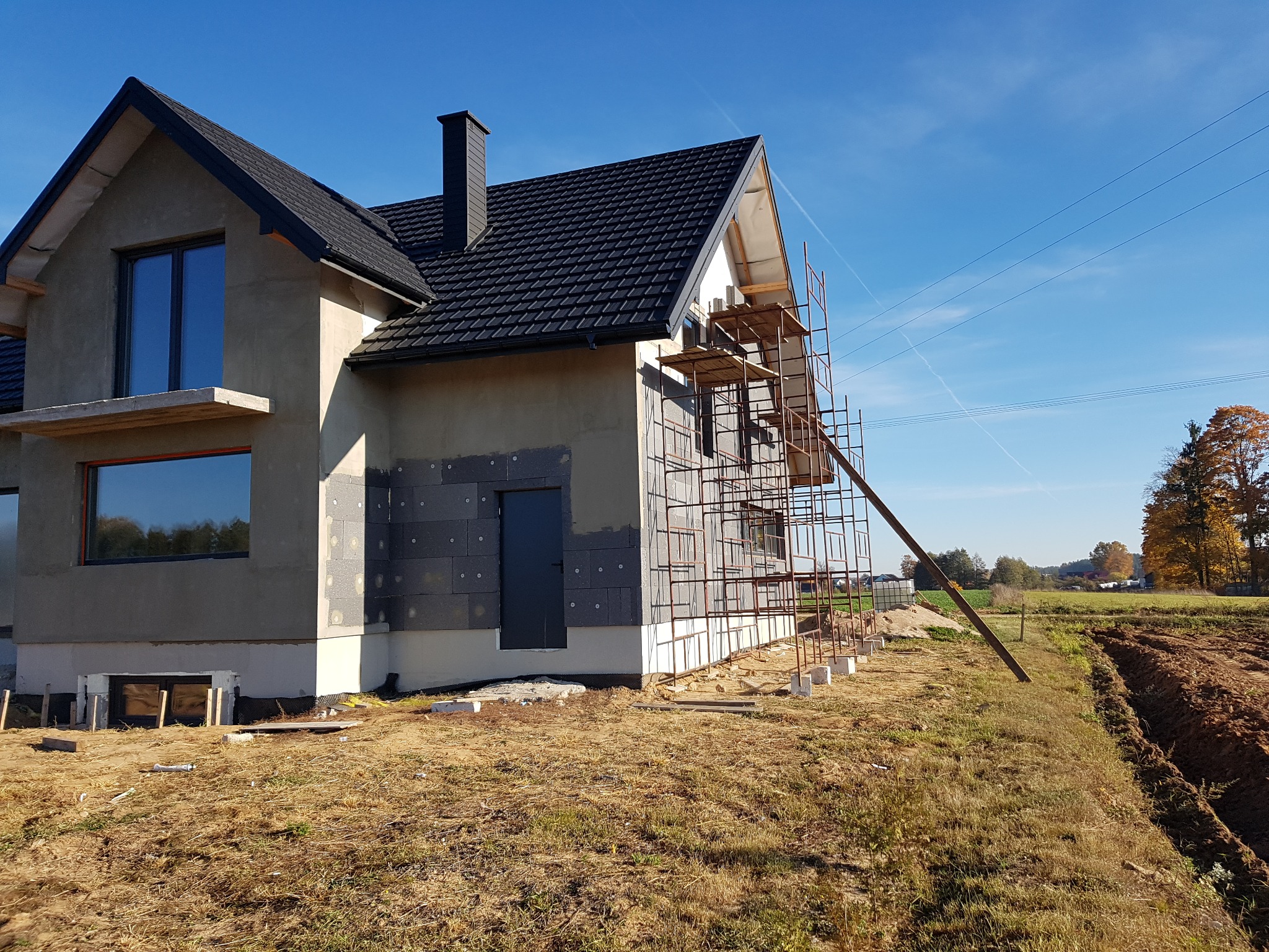 Dom w trakcie ocieplania, widoczne szare bloczki izolacyjne na ścianie, rusztowanie i wykop w ziemi, błękitne niebo w tle.