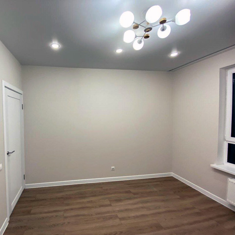 Pomalowane na beżowo puste wnętrze z drewnianą podłogą, nowoczesną lampą sufitową i białymi listwami przypodłogowymi. Widok na okno i drzwi.