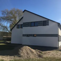 Nowoczesny dom z jasną elewacją, ciemnymi oknami i poziomymi szarymi pasami, widoczny pryzmat piasku i drzewo w tle, błękitne niebo.