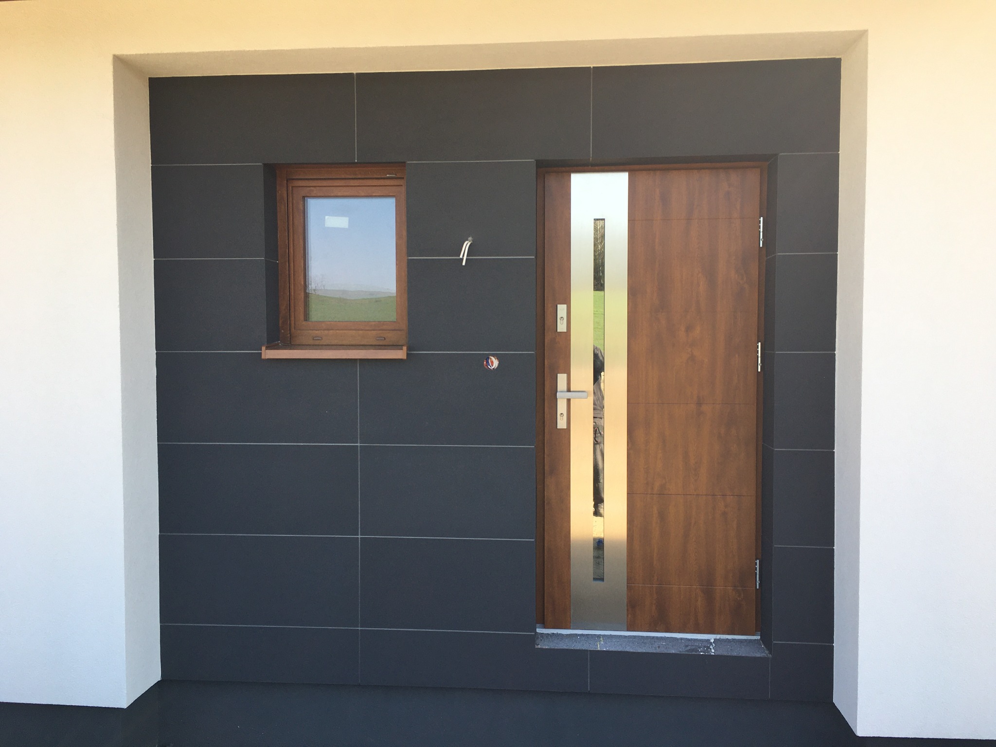 Nowoczesna elewacja domu z antracytowymi płytkami, drewnianą stolarką okienną i drzwi wejściowe z elementami stali nierdzewnej.