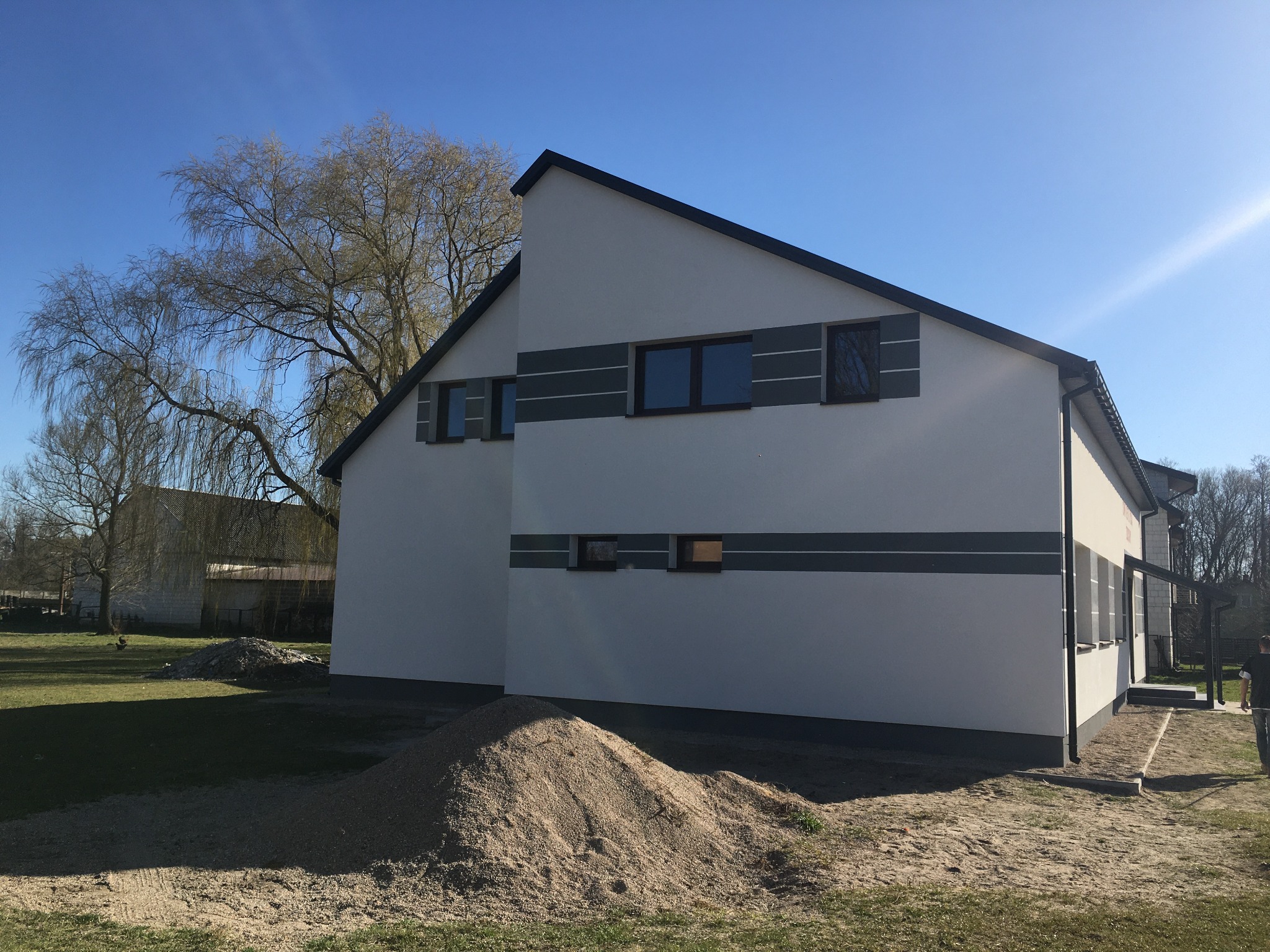 Nowoczesny dom z jasną elewacją, ciemnymi oknami i poziomymi szarymi pasami, widoczny pryzmat piasku i drzewo w tle, błękitne niebo.