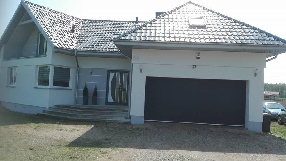 Nowoczesny dom jednorodzinny z szarą elewacją, ciemną bramą garażową i dachem z szarej dachówki, widok z zewnątrz w słoneczny dzień.