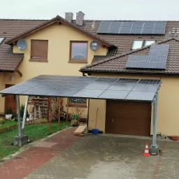 Instalacja paneli słonecznych nad wjazdem do garażu, widoczne panele na dachu domu jednorodzinnego z brązową dachówką i anteną satelitarną.