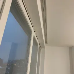 Wnętrze pomieszczenia z oknem, widoczne fragmenty ramy okiennej i gładka, biała ściana oraz sufit z zamontowaną szyną karniszową. Za oknem widok na oświetlone budynki w oddali.