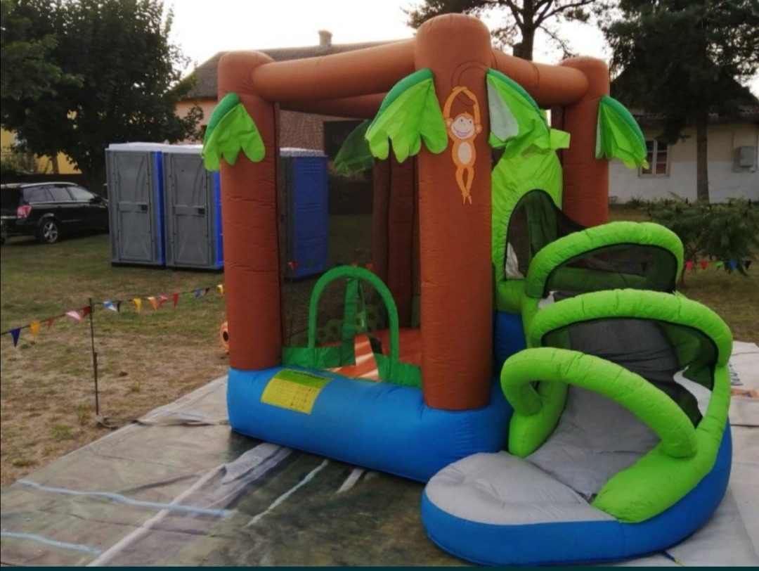 Nadmuchiwaniec w kształcie drzewa z małpką, obok toalety przenośne i girlanda z trójkątnych flag na trawniku.