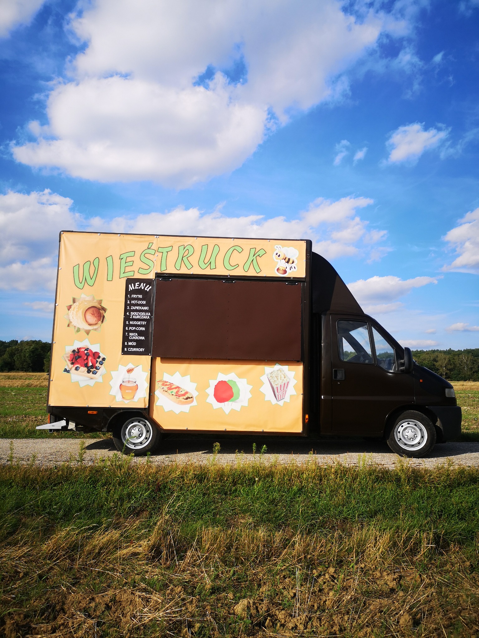 Food truck 'Wieśtruck' z menu wypisanym na boku, oferujący frytki, hot-dogi, zapiekanki i inne przekąski, zaparkowany na skraju pola w okolicach Połańca, pod błękitnym niebem z białymi chmurami.