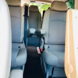 Wnętrze przestronnego busa z jasnymi skórzanymi fotelami i regulowanymi podłokietnikami, widok z perspektywy pasażera z przodu, z pasami bezpieczeństwa i czarną wykładziną podłogową.