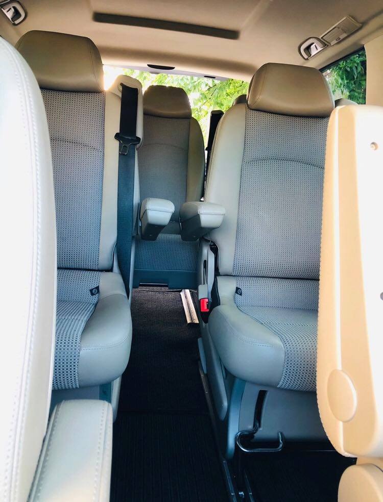 Wnętrze przestronnego busa z jasnymi skórzanymi fotelami i regulowanymi podłokietnikami, widok z perspektywy pasażera z przodu, z pasami bezpieczeństwa i czarną wykładziną podłogową.