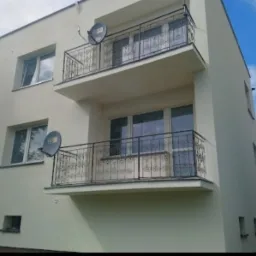 Odnowiona elewacja budynku dwupiętrowego z dwoma balkonami z ozdobnymi, metalowymi balustradami, widoczne okna z roletami oraz satelity na balkonach, jasne niebo z chmurami w tle.