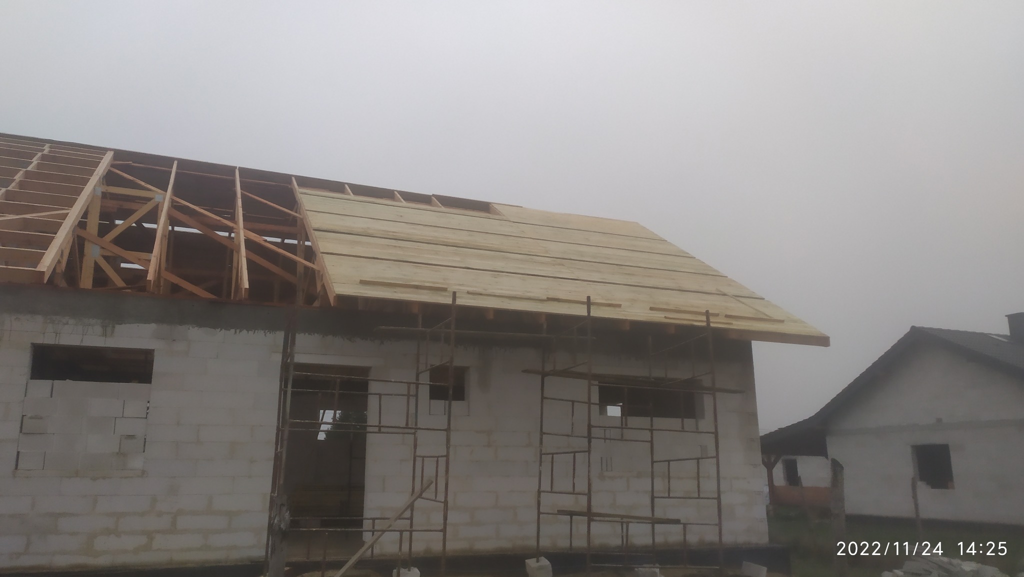 Budowa domu w trakcie prac dekarskich: widoczna drewniana konstrukcja dachu z częściowo położonym deskowaniem, budynek z białego bloczka i rusztowania, mglista pogoda.