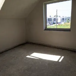 Surowe wnętrze poddasza z oknem i świeżo nałożonym tynkiem na ścianach, widoczny fragment podłogi w stanie surowym, promień słońca wpadający przez okno oświetla podłogę.