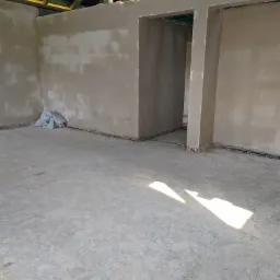 Surowe wnętrze w trakcie prac wykończeniowych, ściany pokryte świeżym tynkiem, widoczne drewniane elementy konstrukcji dachu i cień padający na podłogę.