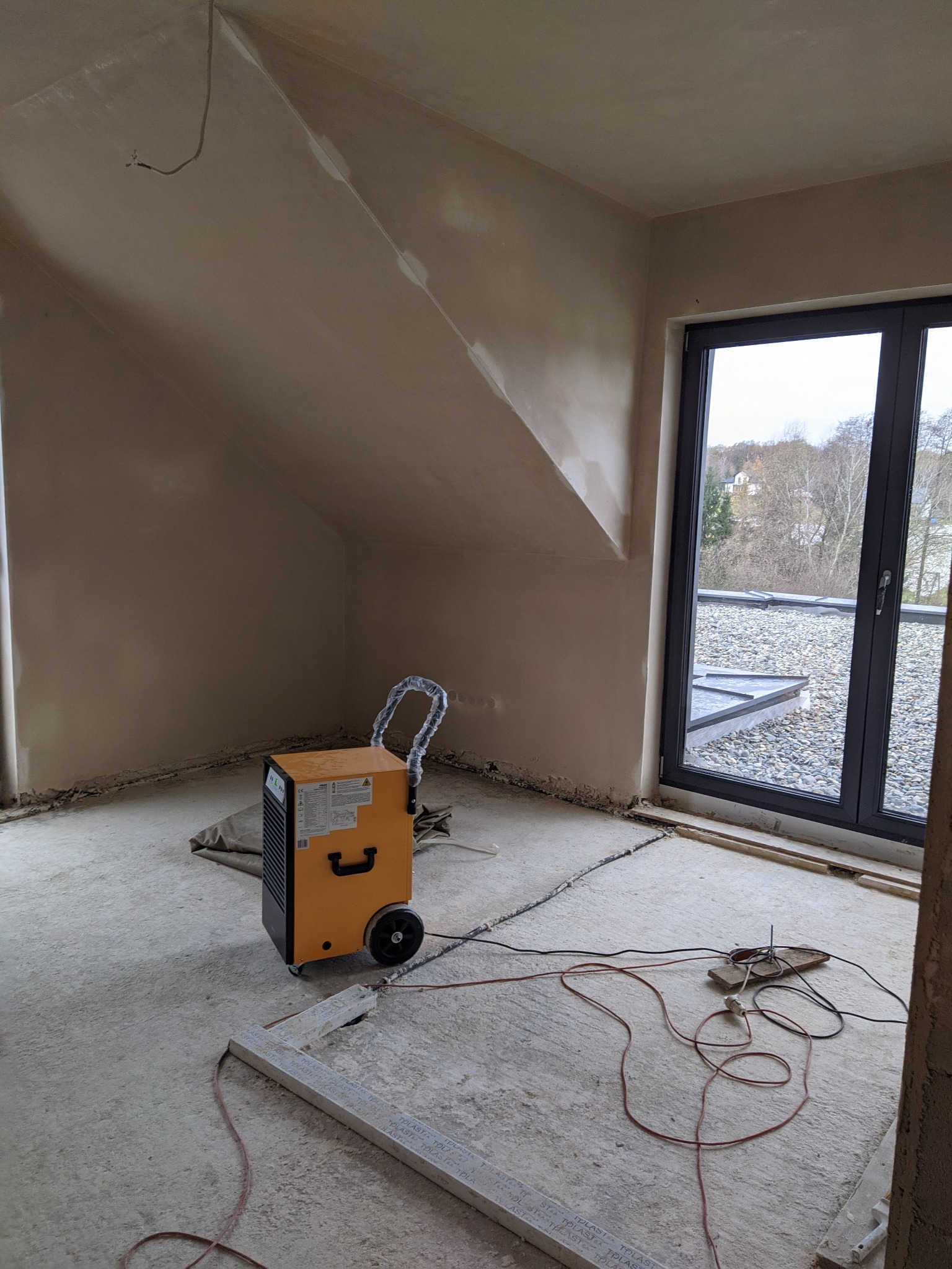 Pokój na poddaszu w trakcie prac wykończeniowych, ściany pokryte świeżym tynkiem, widoczny osuszacz powietrza, przewody elektryczne na podłodze i okno z widokiem na dach pokryty żwirem.