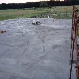 Świeżo wylana płyta fundamentowa z wystającymi prętami zbrojeniowymi, widok z góry na tle zielonego pola i lasu.