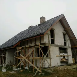 Surowy stan domu z bloczków, z ciemną dachówką, drewnianymi rusztowaniami i kominem z cegły, w trakcie budowy, na tle pochmurnego nieba.