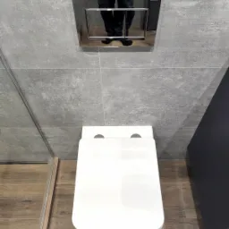 Nowoczesna, podwieszana toaleta z białą deską i chromowanym przyciskiem spłukiwania Geberit, zamontowana na szarej, płytkowej ścianie, z widocznym odbiciem osoby w przycisku.