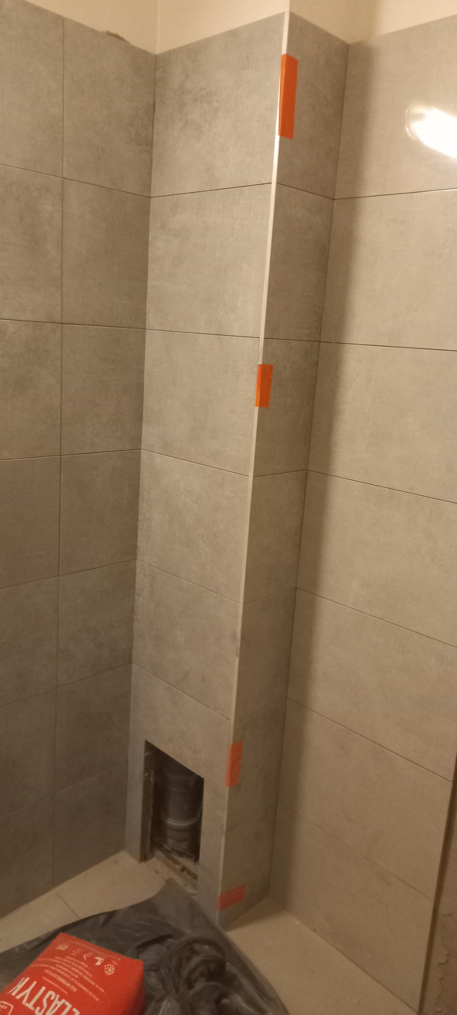 Szara, pozioma glazura w rogu łazienki, z pomarańczową taśmą maskującą na pionowym elemencie wykończeniowym i widocznym fragmentem rury w otworze, sugerująca etap prac wykończeniowych.