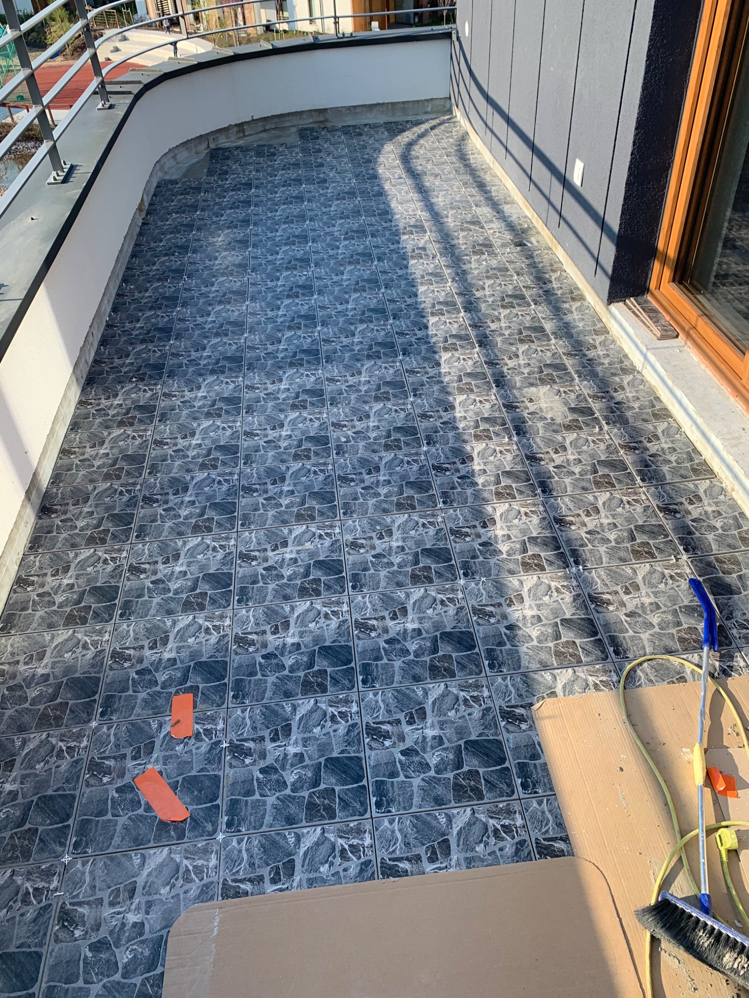 Wyłożony szarymi płytkami balkon z widocznymi krzyżykami dystansowymi i narzędziami do czyszczenia, w tle barierka i elewacja budynku.