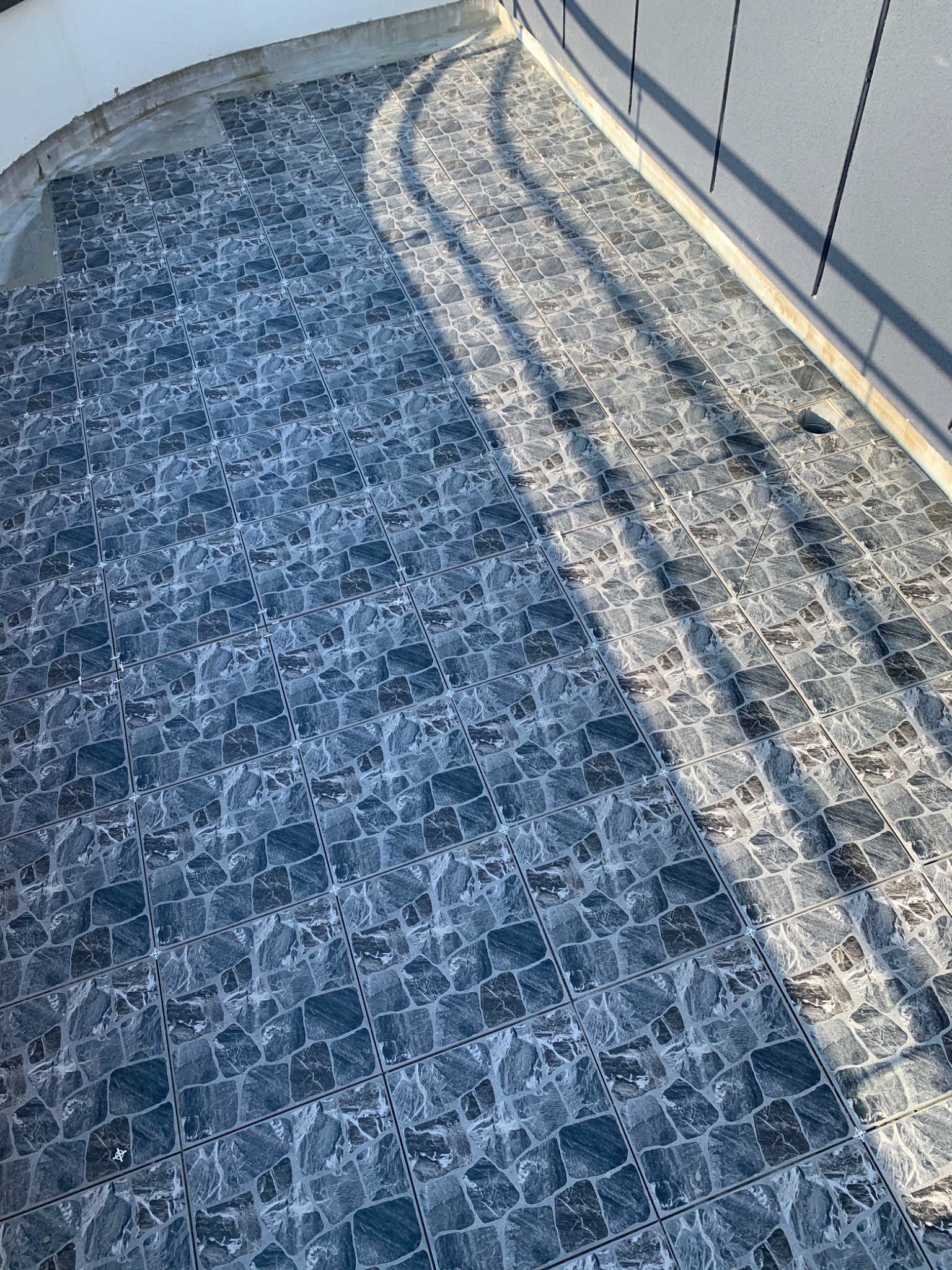 Ułożona, szara, wzorzysta terakota na zewnątrz budynku, widoczne cienie rzucane przez balustradę, fugi z krzyżykami dystansowymi.