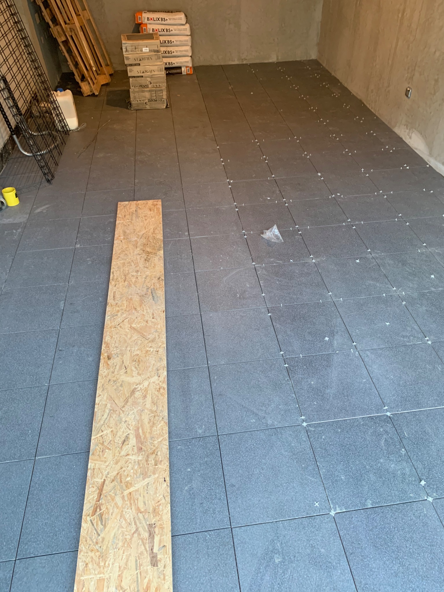 Układanie ciemnoszarych płytek podłogowych z krzyżykami dystansowymi w trakcie prac w Warszawie, widoczna płyta OSB i worki z zaprawą w tle.