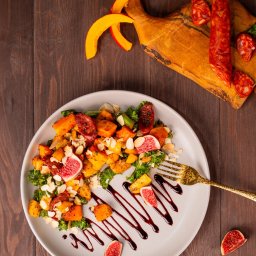 Fotografia kulinarna Kalbarczyk - Jesienna sałatka z pieczonej dyni, chorizo, fig, jarmużu i płatków migdałów, polana sosem balsamicznym, serwowana na szarym talerzu z pozłacanym widelcem na drewnianym stole, z deską do krojenia...