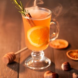 Fotografia kulinarna Kalbarczyk - Szklanka parującego napoju z pomarańczą, cynamonem i gałązką rozmarynu na drewnianym stole z orzechami i suszonymi pomarańczami, obok nabierak do miodu.