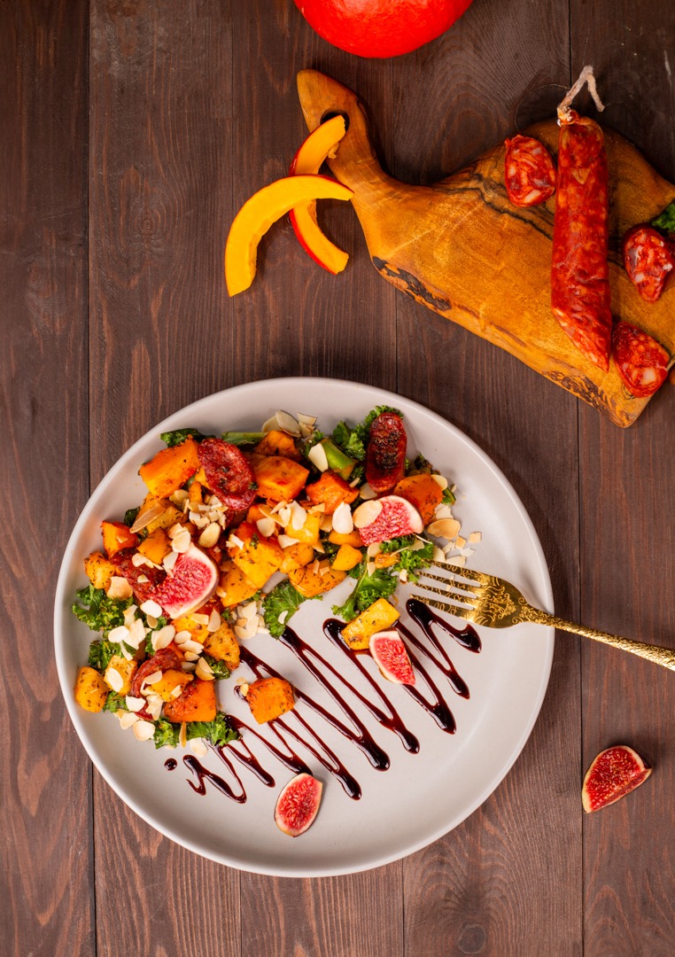 Jesienna sałatka z pieczonej dyni, chorizo, fig, jarmużu i płatków migdałów, polana sosem balsamicznym, serwowana na szarym talerzu z pozłacanym widelcem na drewnianym stole, z deską do krojenia...