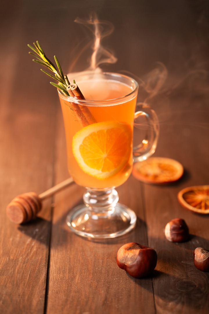 Szklanka parującego napoju z pomarańczą, cynamonem i gałązką rozmarynu na drewnianym stole z orzechami i suszonymi pomarańczami, obok nabierak do miodu.