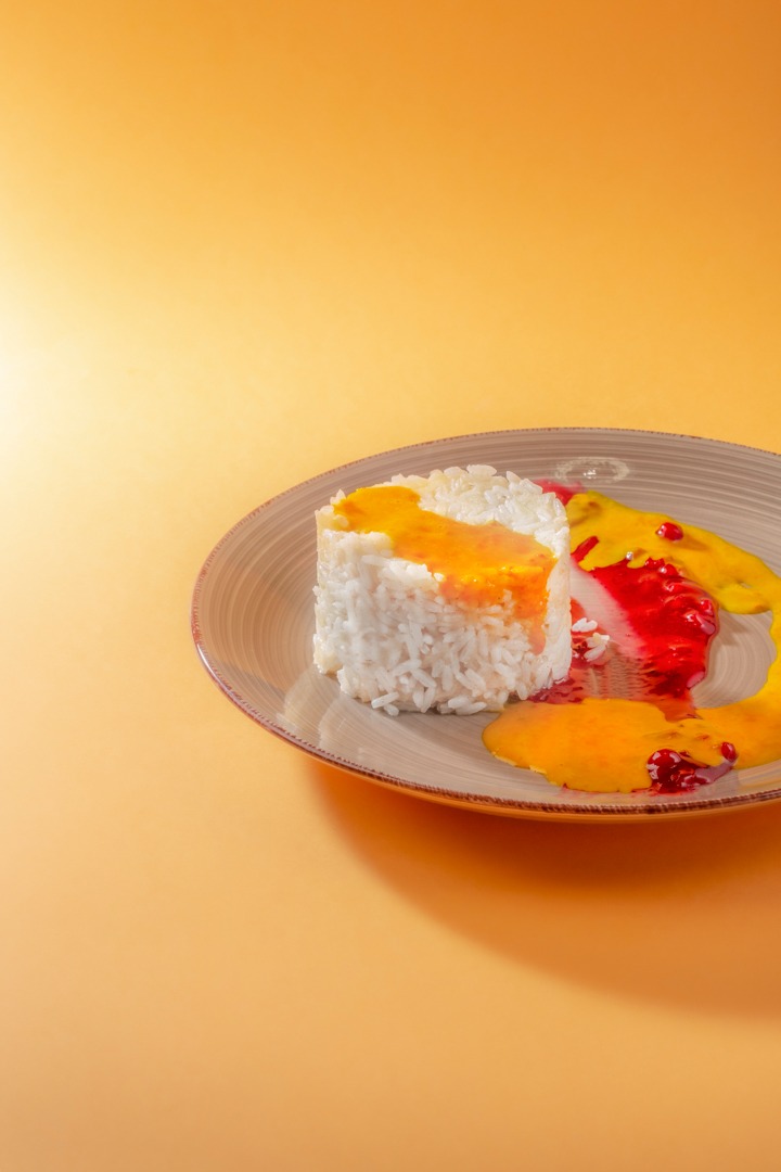 Ułożona w walec porcja białego ryżu polana pomarańczowym sosem, obok kleksy czerwonego sosu z owocami na talerzu z szarą obwódką, na pomarańczowym tle.