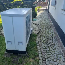 Biała pompa ciepła typu monoblok ustawiona na betonowych bloczkach przy ścianie domu, obok wyłożony kostką brukową chodnik i trawnik z drzewem w tle.