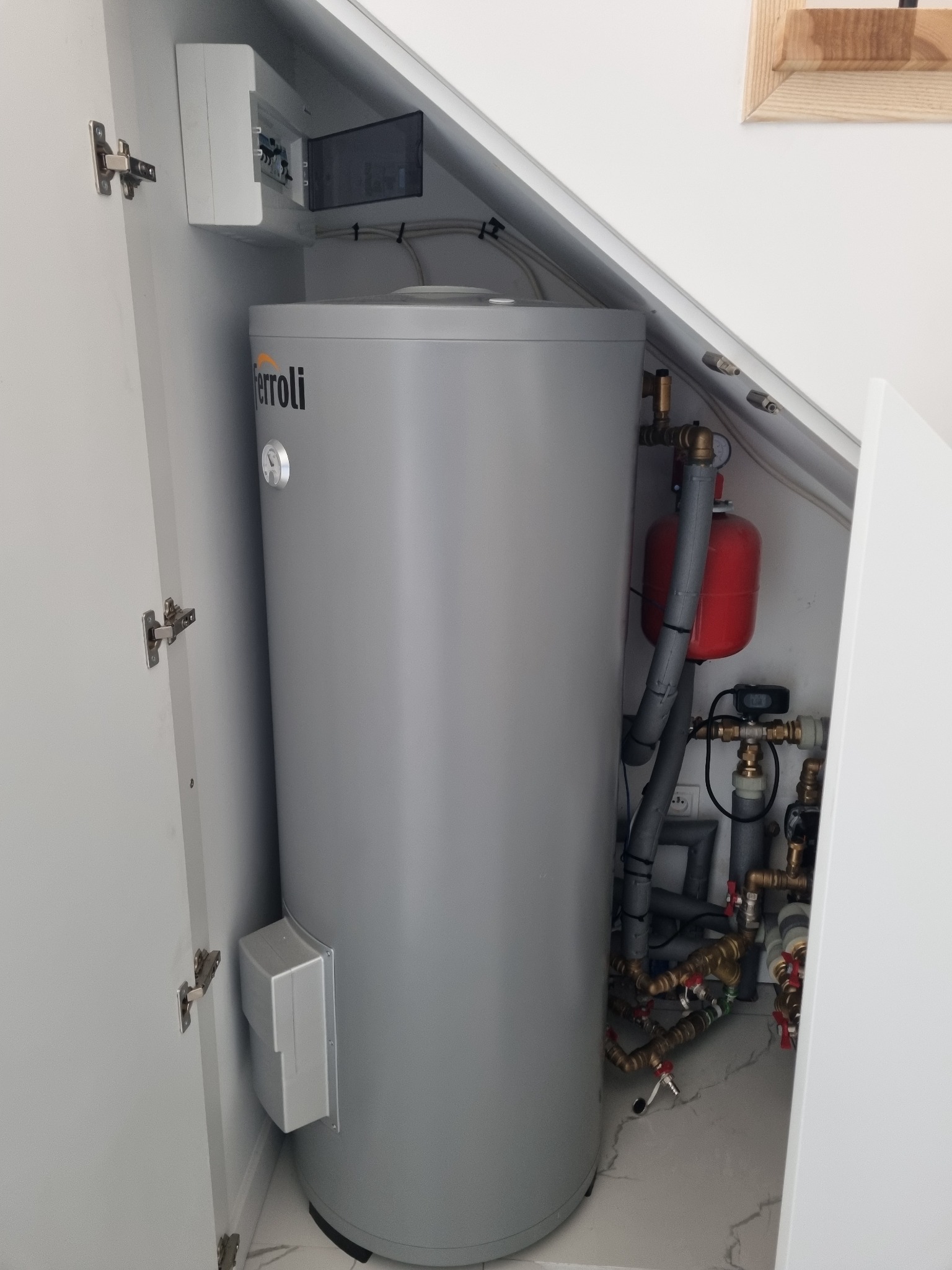 Szary zasobnik ciepłej wody Ferroli w zabudowie szafkowej, widoczne rury, zawory i osprzęt instalacyjny.