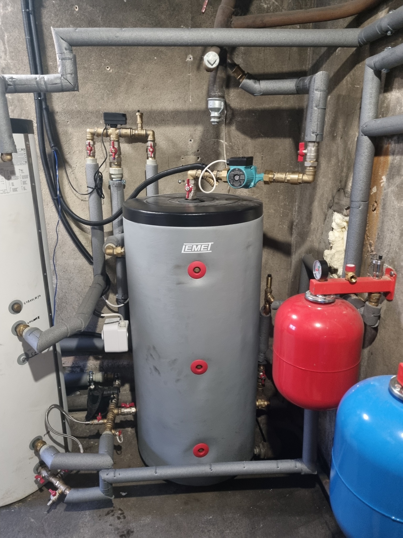 Szara instalacja centralnego ogrzewania z zbiornikiem buforowym, czerwonym naczyniem przeponowym i niebieskim zbiornikiem, rury zaizolowane szarą pianką, widoczne zawory i manometr.