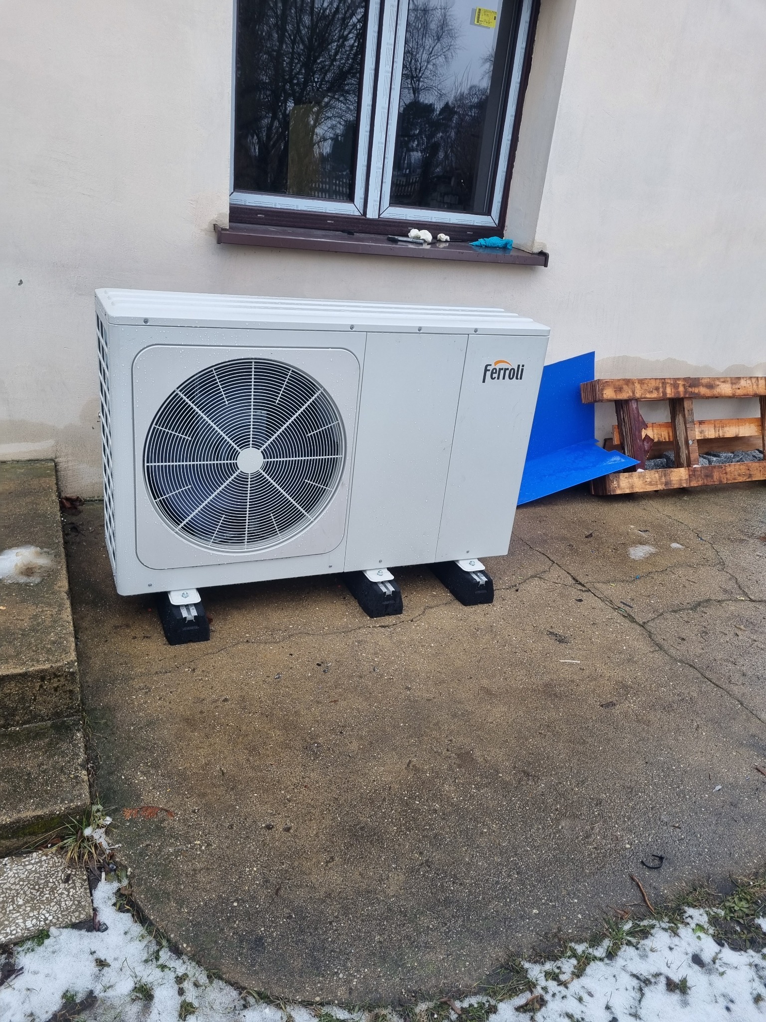 Biała pompa ciepła marki Ferroli zamontowana na betonowym podłożu przy ścianie budynku z oknem, widoczne ślady śniegu.