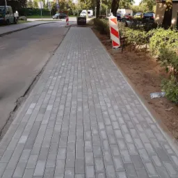 Świeżo ułożona, szara kostka brukowa wzdłuż drogi, z widocznymi znakami ostrzegawczymi i ziemią po bokach, sugerująca trwające prace wykończeniowe.